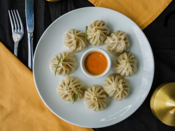 A White Plate With Eight Dumplings Arranged In A Circle Around A Small Bowl Of Orange Dipping Sauce, Placed On A Table With Yellow And Black Napkins.