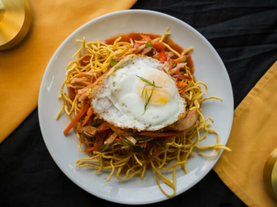 A White Plate Of Fried Noodles Topped With Vegetables, Shredded Chicken, And A Sunny Side Up Egg, Placed On A Black And Yellow Cloth Background.