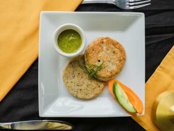 A White Plate With Two Patties, A Small Cup Of Green Sauce, And Garnished With A Slice Of Carrot And Cucumber, On A Table With Yellow And Black Tablecloths.