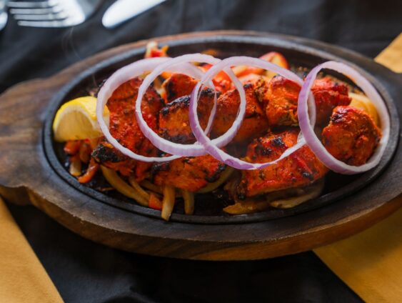 A Sizzling Platter Of Grilled Chicken Kebabs Garnished With Onion Rings And Accompanied By Lemon Slices, On A Dark Tablecloth With A Fork, Knife, And Gold Decor Nearby.