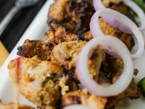 Grilled Chicken Skewers Garnished With Onion Rings On A White Plate, Accompanied By Greens. A Fork Is Visible In The Background.