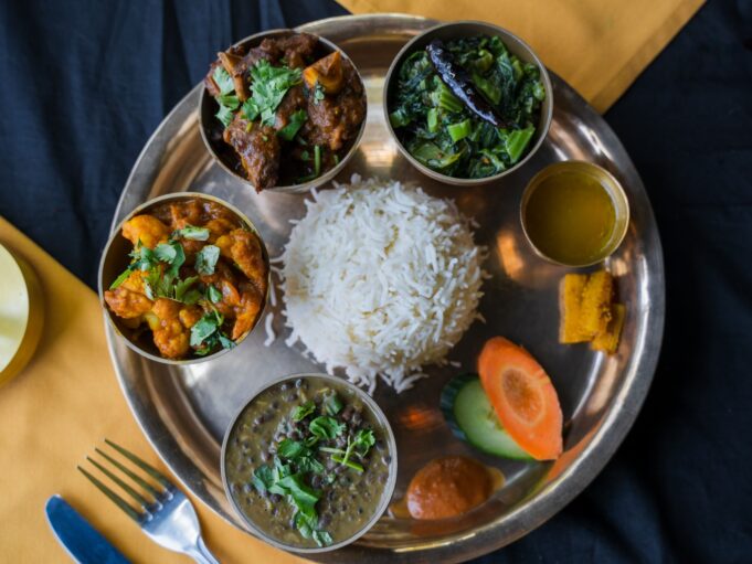 A Traditional Indian Meal Served On A Metal Platter, Featuring Rice, Assorted Curries, Lentils, Greens, Sliced Carrots, Cucumber, And A Small Dish Of Sauce.